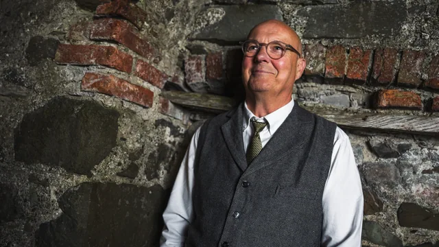 A musician in a waistcoat photographed against a textured stone wall
